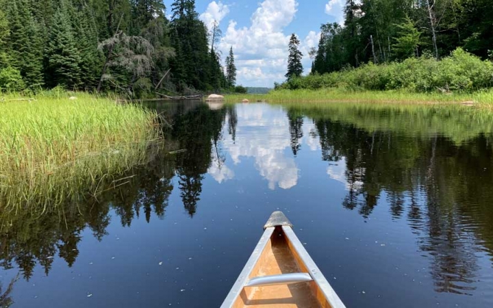 In the foreground, the tip of a canoe appears above very calm water, reflecting the grasses and trees on the shore, and the blue sky and clouds above. 